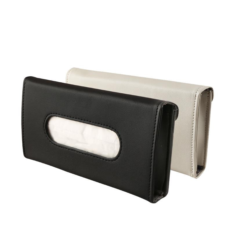 Two car visor tissue holders on white background — black variant in front and beige variant behind — both showing the oval tissue slot on the front face