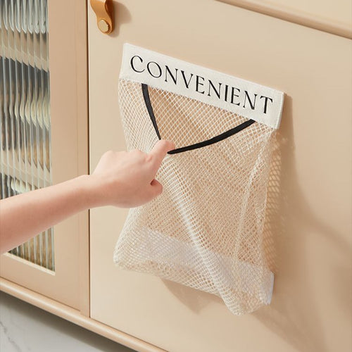 Hand pulling a plastic bag from a beige wall-mounted mesh storage bag organizer labeled CONVENIENT, hung on a cream kitchen cabinet door