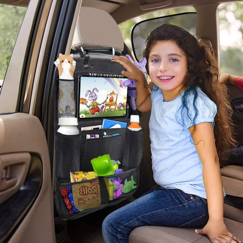 Young girl in the back seat of a car using a black multi-pocket backseat organizer with clear tablet holder showing a cartoon, filled with toys, bottles, and snacks