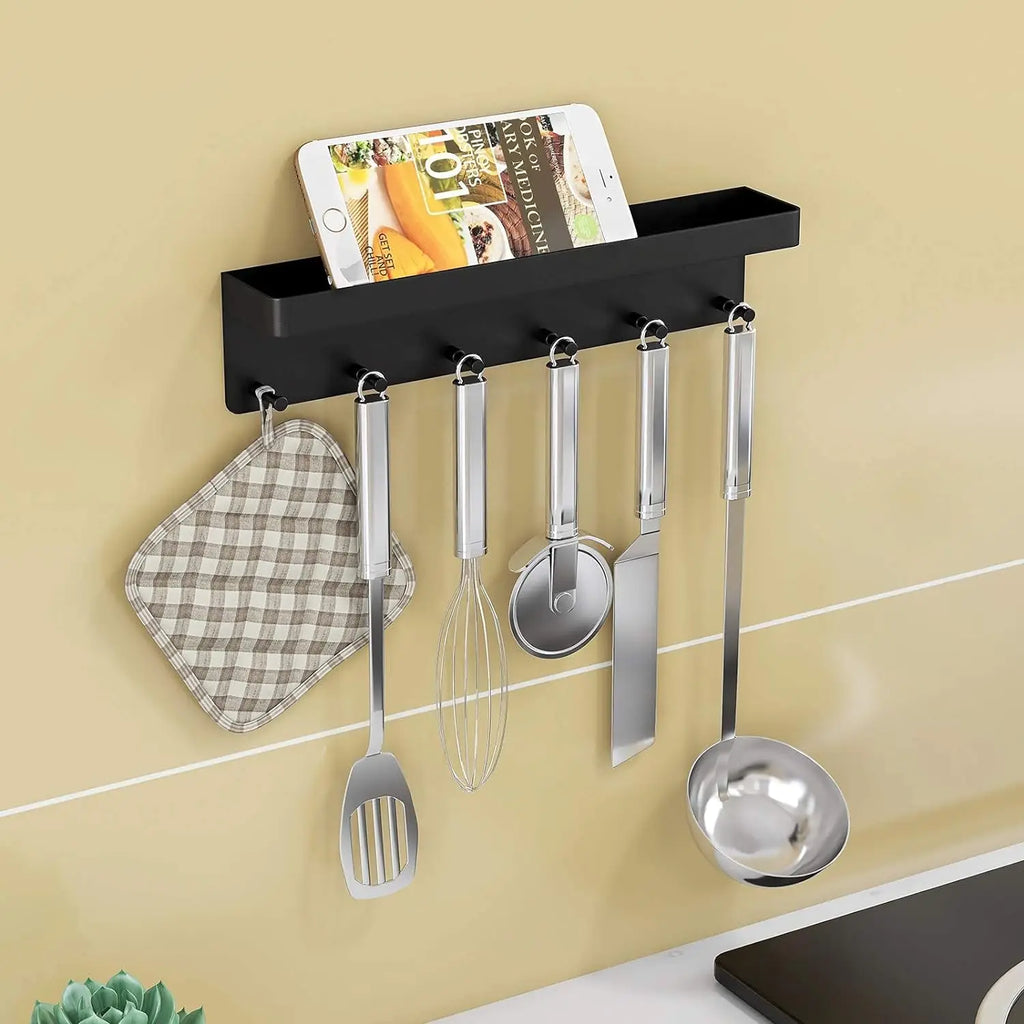Black wall-mounted key and storage shelf used in a kitchen on a yellow tiled wall, top shelf holding a smartphone and cookbook, hooks below holding a checkered pot holder, whisk, pizza cutter, spatula, and ladle