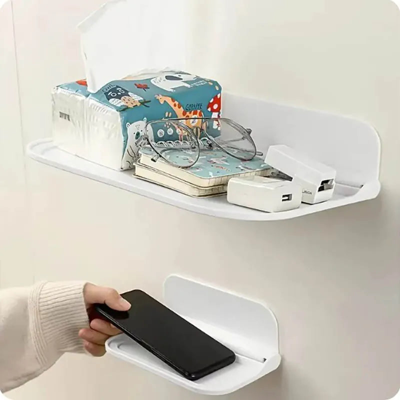 Two white foldable wall-mounted shelves on a white wall — large shelf holding a tissue box, glasses, notebook, and USB hub above; hand placing a smartphone on the small shelf below