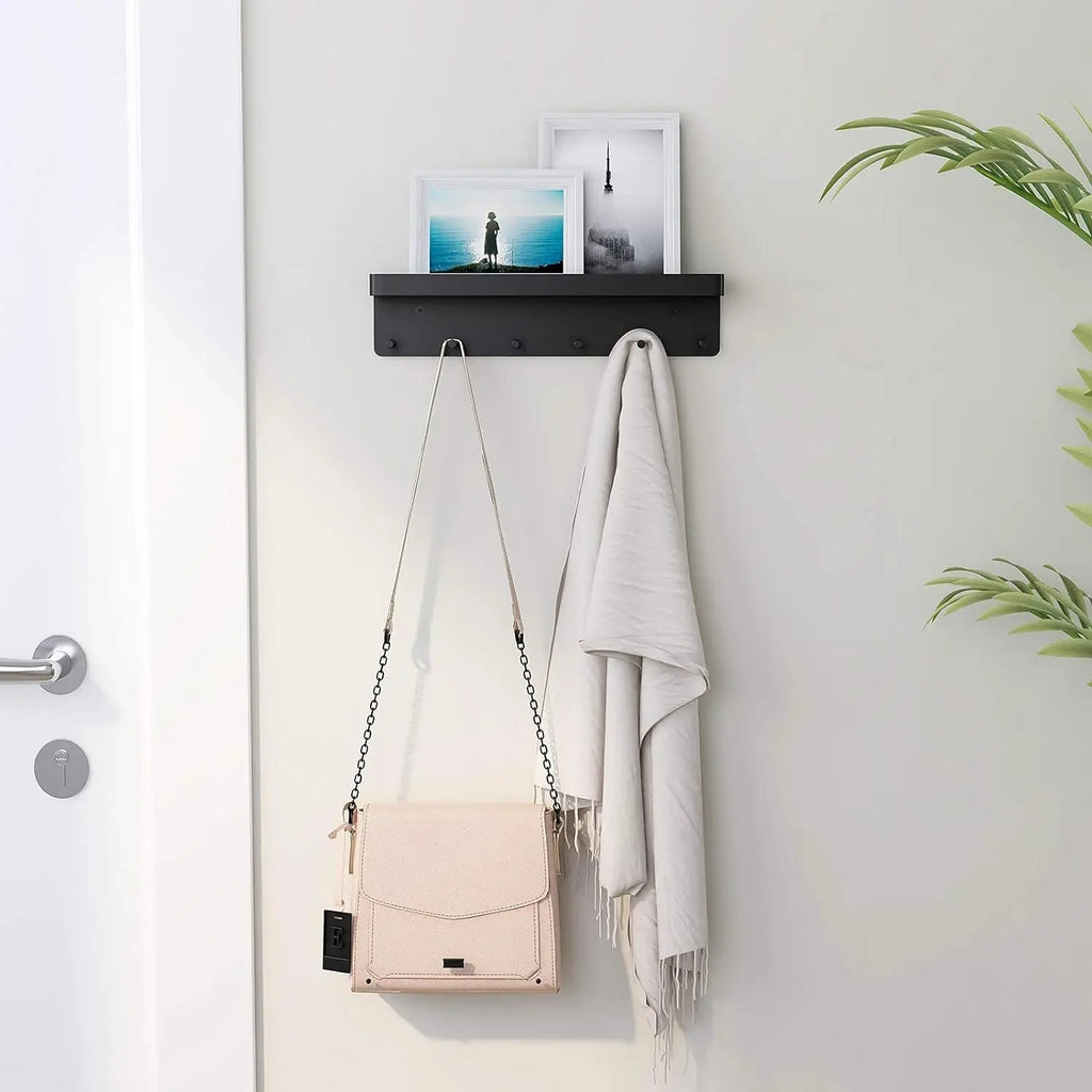 Black wall-mounted key and mail organizer shelf mounted beside a front door on a light gray wall, holding photo frames on the top shelf with a crossbody bag and white scarf hanging from the hooks below