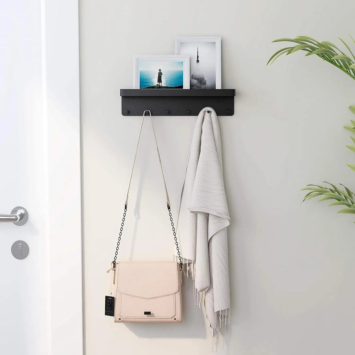 Black wall-mounted key and mail organizer shelf mounted beside a front door on a light gray wall, holding photo frames on the top shelf with a crossbody bag and white scarf hanging from the hooks below