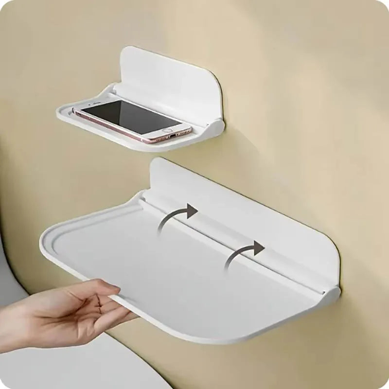 Two white foldable wall-mounted shelves on a beige wall — small shelf with a smartphone resting on it above, and a hand unfolding the larger shelf below, showing the fold-out mechanism with directional arrows